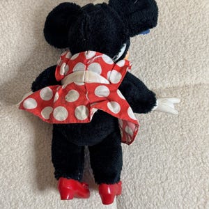 May include: A plush Minnie Mouse doll, viewed from the back, wearing a red dress with white polka dots and red shoes. The doll has black fur, large black ears, and white gloves. The doll is posed with one arm extended.