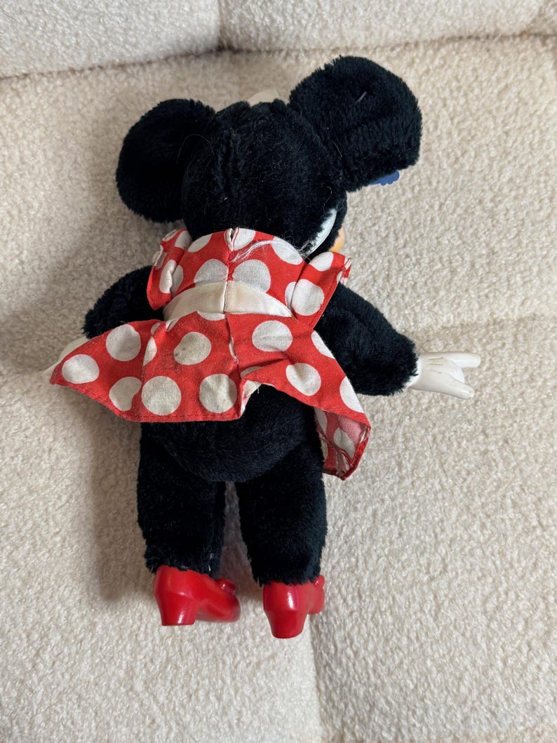 May include: A plush Minnie Mouse doll, viewed from the back, wearing a red dress with white polka dots and red shoes. The doll has black fur, large black ears, and white gloves. The doll is posed with one arm extended.
