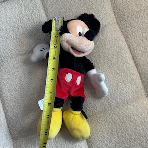May include: A Mickey Mouse plush toy, approximately 12 inches tall, is shown. The plush features black fur, a tan face, red shorts with white buttons, yellow shoes, and white gloves. A yellow measuring tape is next to the toy.