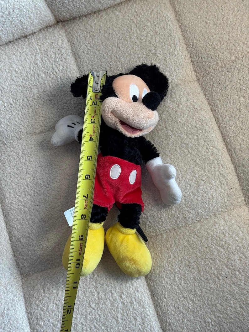 May include: A Mickey Mouse plush toy, approximately 12 inches tall, is shown. The plush features black fur, a tan face, red shorts with white buttons, yellow shoes, and white gloves. A yellow measuring tape is next to the toy.