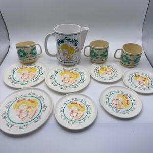May include: A vintage Cabbage Patch Kids themed dining set. The set includes a white pitcher, three mugs, and several plates. The plates and mugs feature a repeating floral pattern and a Cabbage Patch Kids graphic. The pitcher has the Cabbage Patch Kids logo.