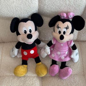 May include: Two plush Disney character dolls, Mickey Mouse and Minnie Mouse, are displayed. Mickey wears red shorts with white buttons, yellow shoes, and black ears. Minnie is in a pink dress with white polka dots, pink shoes, and a pink bow.
