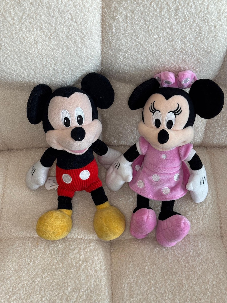May include: Two plush Disney character dolls, Mickey Mouse and Minnie Mouse, are displayed. Mickey wears red shorts with white buttons, yellow shoes, and black ears. Minnie is in a pink dress with white polka dots, pink shoes, and a pink bow.