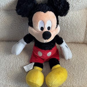 May include: A plush Mickey Mouse toy with black fur, large black ears, and a tan face. He wears red shorts with white buttons, yellow shoes, and white gloves. The toy is sitting on a textured, off-white surface.