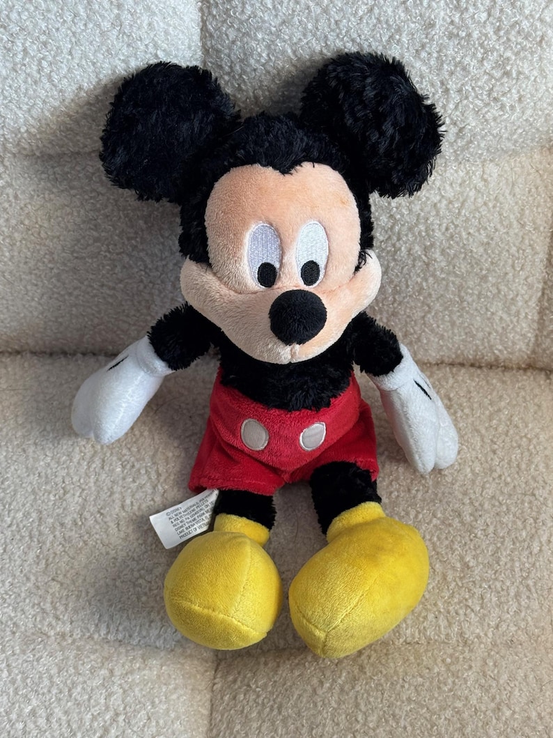 May include: A plush Mickey Mouse toy with black fur, large black ears, and a tan face. He wears red shorts with white buttons, yellow shoes, and white gloves. The toy is sitting on a textured, off-white surface.
