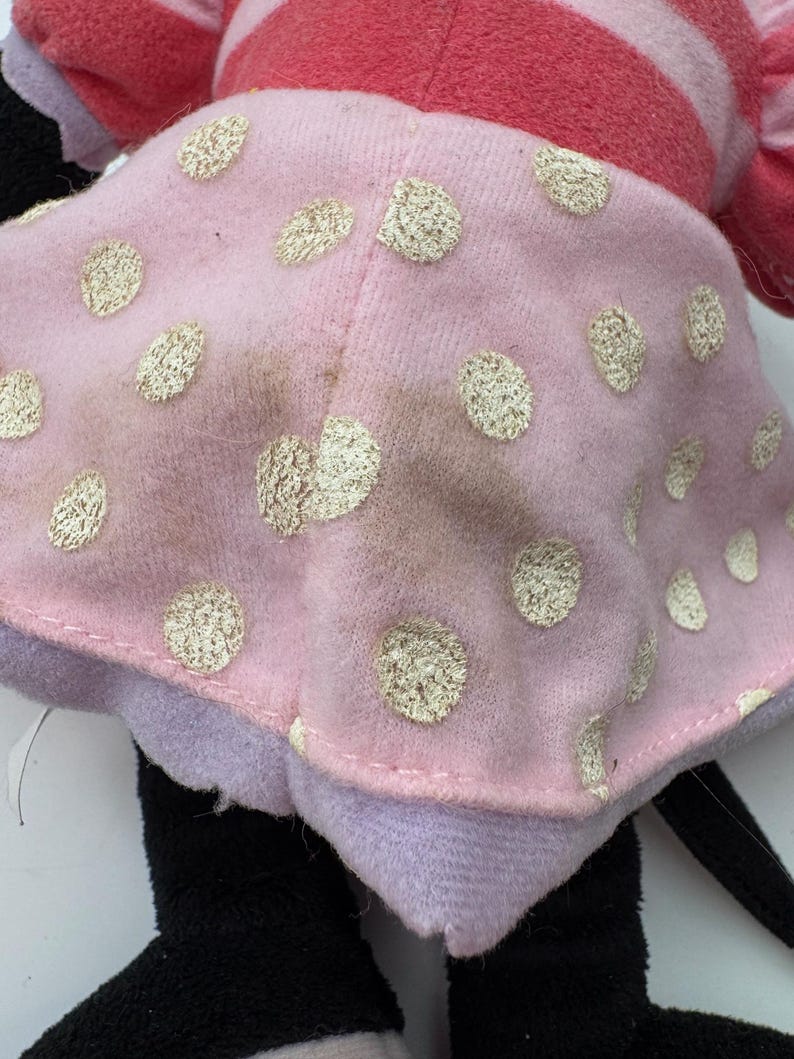 May include: A plush doll featuring a pink dress with gold polka dots. The doll has a red and pink striped top, black legs, and a black tail. The doll's dress has some discoloration.