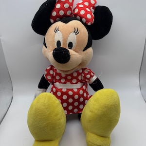May include: A plush Minnie Mouse doll, featuring a red dress with white polka dots and a matching bow. The doll has black ears, a black nose, and yellow feet. The doll is sitting upright.