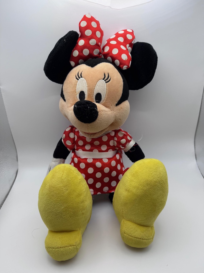 May include: A plush Minnie Mouse doll, featuring a red dress with white polka dots and a matching bow. The doll has black ears, a black nose, and yellow feet. The doll is sitting upright.