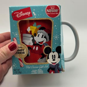 May include: A Disney Hot Cocoa Gift Set featuring Mickey Mouse. The set includes a mug, candy stirrer, and hot cocoa mix. The box is light blue with a gold border and snowflake design. The net weight is 28g.