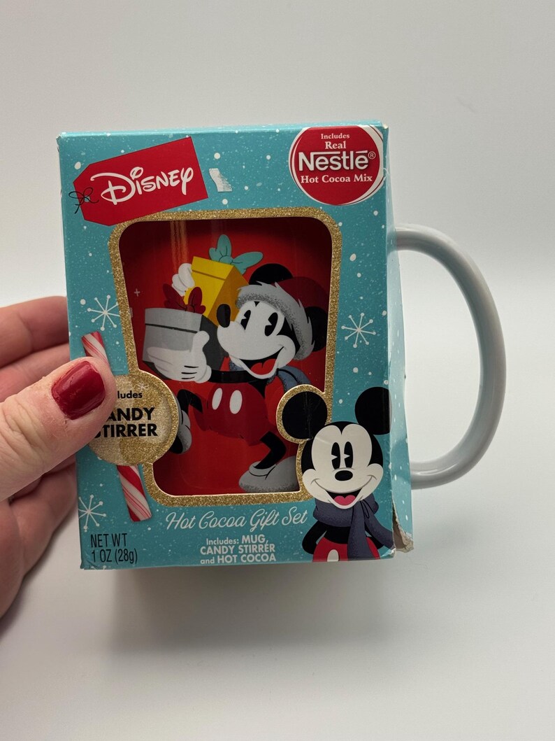May include: A Disney Hot Cocoa Gift Set featuring Mickey Mouse. The set includes a mug, candy stirrer, and hot cocoa mix. The box is light blue with a gold border and snowflake design. The net weight is 28g.