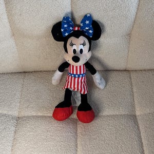 May include: A Minnie Mouse soft toy dressed in patriotic attire. The toy features a blue bow with white stars, a red, white, and blue striped dress, and red shoes. The black ears and face of the character are visible.