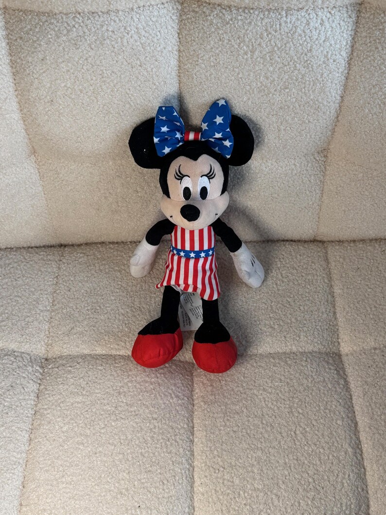 May include: A Minnie Mouse soft toy dressed in patriotic attire. The toy features a blue bow with white stars, a red, white, and blue striped dress, and red shoes. The black ears and face of the character are visible.