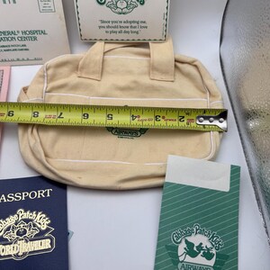 May include: A cream-coloured canvas bag with two handles and white piping, featuring a green Cabbage Patch Kids Airways logo. A yellow measuring tape across the bag shows a width of approximately 21.5 cm. Includes a matching green card and passport.