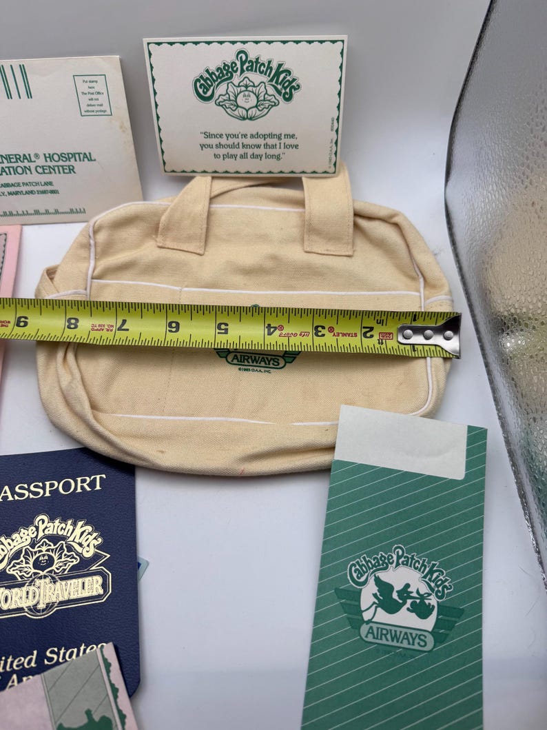 May include: A cream-coloured canvas bag with two handles and white piping, featuring a green Cabbage Patch Kids Airways logo. A yellow measuring tape across the bag shows a width of approximately 21.5 cm. Includes a matching green card and passport.