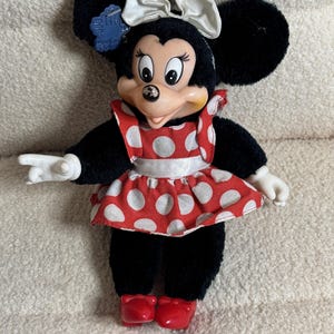 May include: A Minnie Mouse plush doll with black fur, large black ears, and a white bow. She wears a red and white polka dot dress and red shoes. The doll has white gloves and a blue tag.