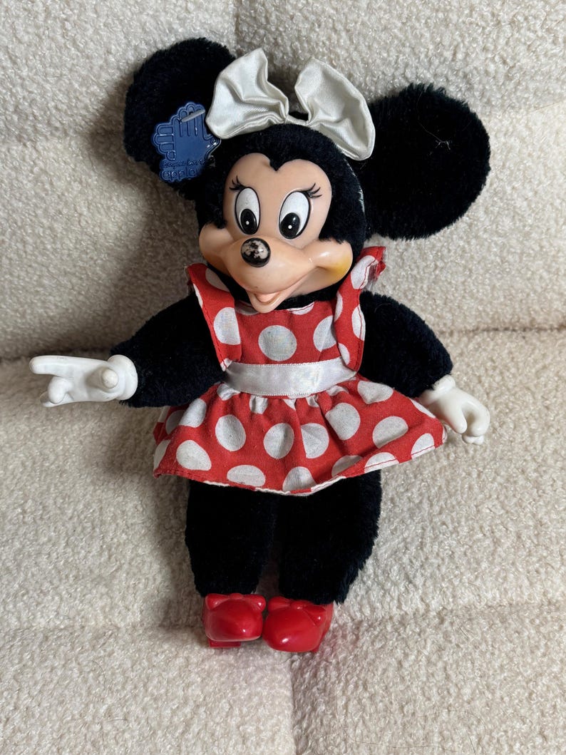 May include: A Minnie Mouse plush doll with black fur, large black ears, and a white bow. She wears a red and white polka dot dress and red shoes. The doll has white gloves and a blue tag.