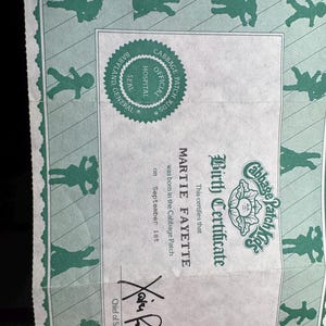 May include: A Cabbage Patch Kids birth certificate printed on light green paper with a dark green border featuring silhouettes of dolls. The document includes the text Birth Certificate, Martie Fayette, September 1st, and a signature.