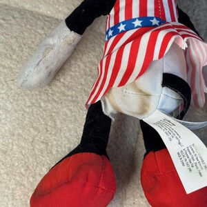 May include: A soft toy character in a red, white, and blue outfit. The outfit is designed like a flag, with red and white stripes and a blue waistband with white stars. The toy has black ears, arms, and legs, and red shoes.