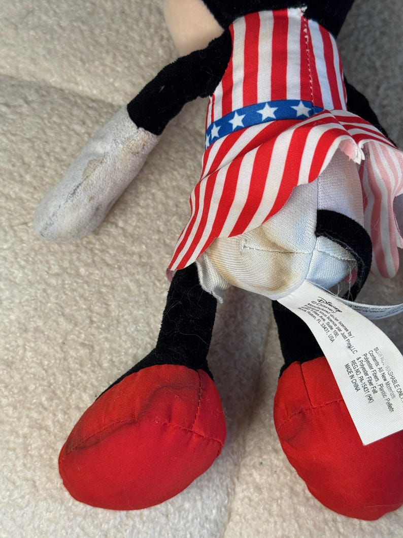 May include: A soft toy character in a red, white, and blue outfit. The outfit is designed like a flag, with red and white stripes and a blue waistband with white stars. The toy has black ears, arms, and legs, and red shoes.