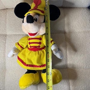 May include: A plush Minnie Mouse doll wearing a yellow marching band uniform with red accents. The doll has black ears, a white face, and is approximately 16 inches tall.