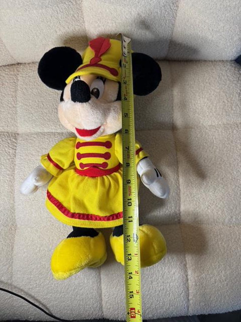 May include: A plush Minnie Mouse doll wearing a yellow marching band uniform with red accents. The doll has black ears, a white face, and is approximately 16 inches tall.