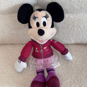 Puede incluir: Mu&ntilde;eca de peluche de Minnie Mouse con una chaqueta granate, un tut&uacute; rosa y calcetines morados. La mu&ntilde;eca tiene orejas negras, cara blanca y nariz negra. La chaqueta tiene cremallera y parches.