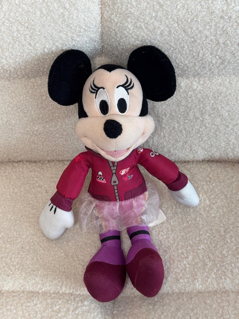 Puede incluir: Mu&ntilde;eca de peluche de Minnie Mouse con una chaqueta granate, un tut&uacute; rosa y calcetines morados. La mu&ntilde;eca tiene orejas negras, cara blanca y nariz negra. La chaqueta tiene cremallera y parches.