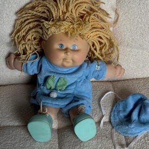 May include: A vintage doll with a round face, blue eyes, and blonde yarn hair. The doll wears a blue outfit with green gingham accents and matching shoes. A blue bonnet with white ties sits nearby. A classic toy.