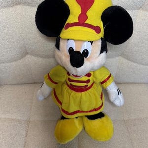 May include: A plush toy of Mickey Mouse wearing a yellow marching band uniform with red accents. The toy has black ears, a yellow hat with a red plume, and white gloves. The outfit includes a yellow tunic and shoes.