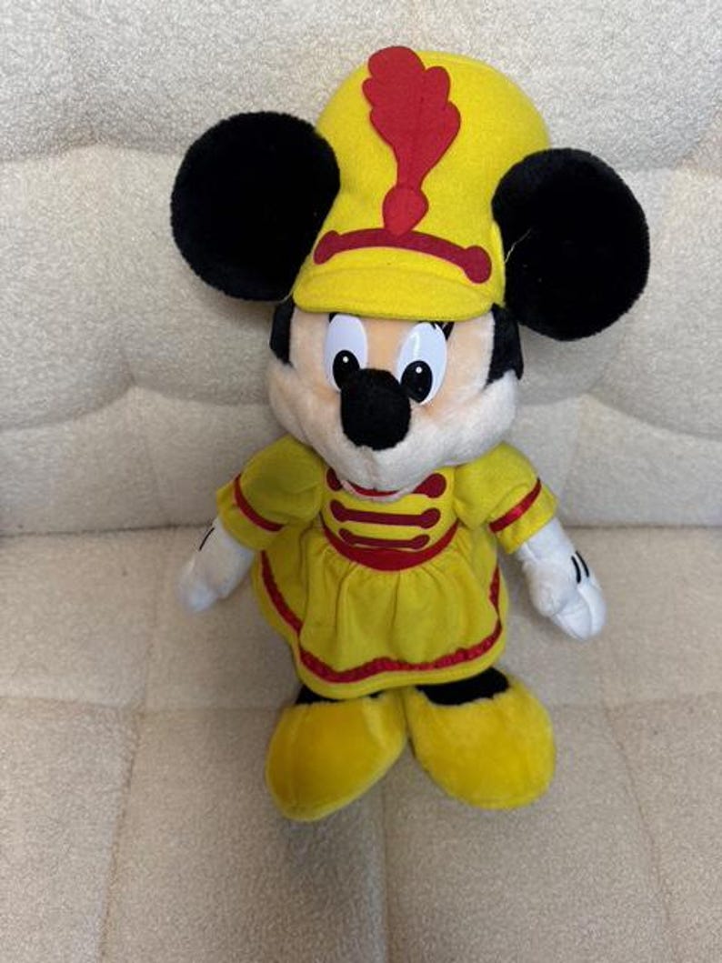 May include: A plush toy of Mickey Mouse wearing a yellow marching band uniform with red accents. The toy has black ears, a yellow hat with a red plume, and white gloves. The outfit includes a yellow tunic and shoes.