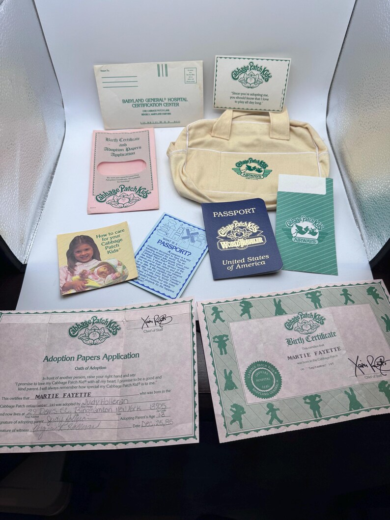 May include: A Cabbage Patch Kids accessory set including a cream-coloured fabric bag, a blue United States of America passport, a pink adoption papers application, a birth certificate for Martie Fayette, and various informational cards and envelopes.