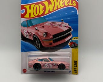 May include: A pink Hot Wheels Datsun 240Z die-cast car with cherry blossom graphics. The car is on a blue and yellow card with the Hot Wheels logo and the text "Guaranteed for Life". The car is 3 inches long.