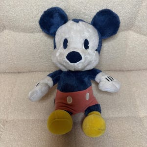 May include: A plush Mickey Mouse toy. It has a white face, dark blue ears and body, red shorts, and yellow feet. The toy has black eyes and a black nose. The hands are white with black lines. The toy is on a beige surface.