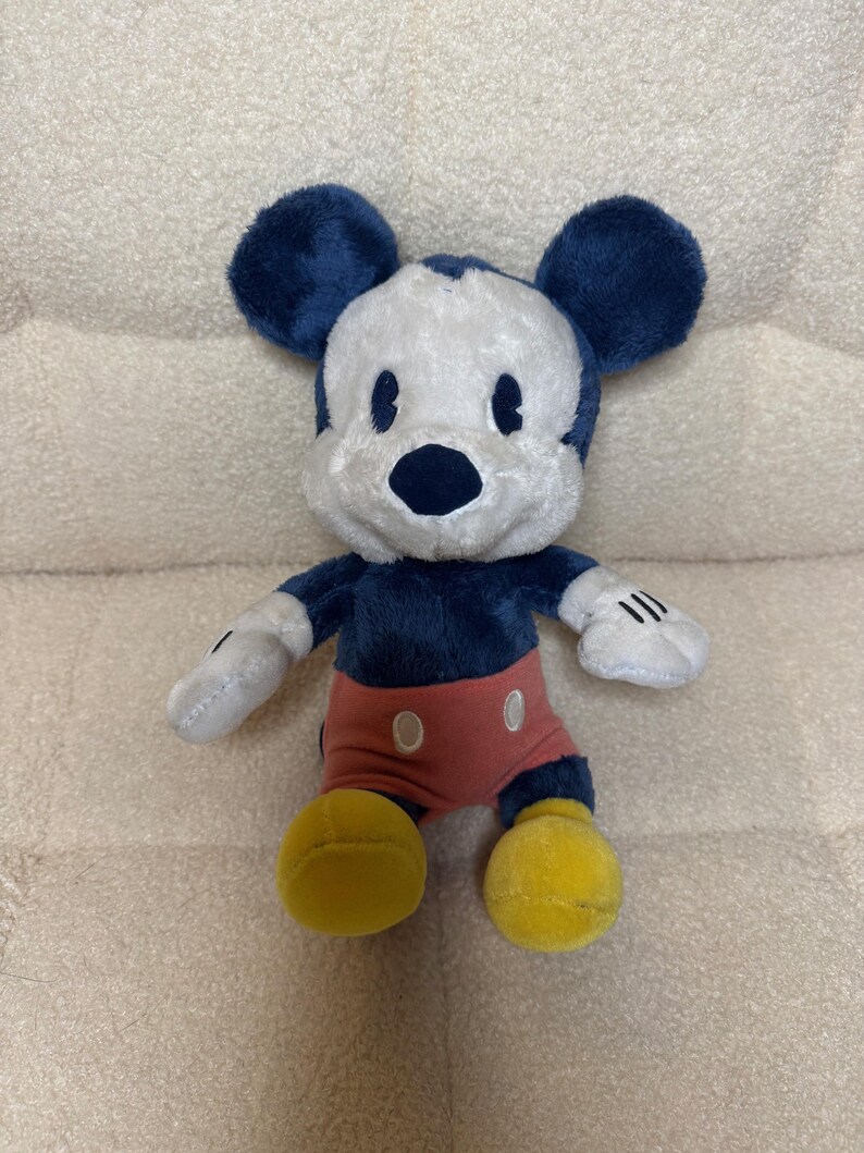 May include: A plush Mickey Mouse toy. It has a white face, dark blue ears and body, red shorts, and yellow feet. The toy has black eyes and a black nose. The hands are white with black lines. The toy is on a beige surface.