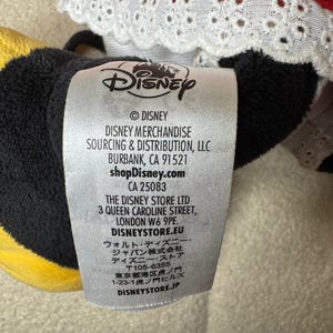 以下が含まれることがあります： ディズニーのぬいぐるみタグのクローズアップ。タグは白地に黒文字で、ディズニーのロゴが入っています。タグには、ディズニーマーチャンダイズソーシング＆ディストリビューションLLCの住所と、ロンドンのディズニー・ストアの住所が記載されています。