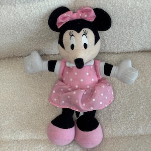 May include: A Minnie Mouse plush toy. The toy has black ears, a black nose, and a pink bow. She is wearing a pink dress with white polka dots and pink shoes. Her arms and hands are white.