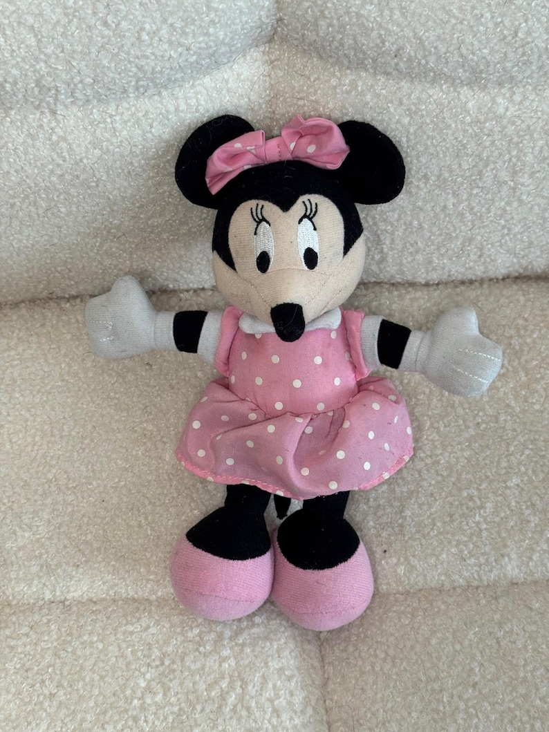 May include: A Minnie Mouse plush toy. The toy has black ears, a black nose, and a pink bow. She is wearing a pink dress with white polka dots and pink shoes. Her arms and hands are white.