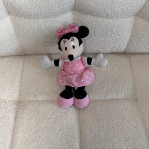 May include: A Minnie Mouse plush doll with a pink polka dot dress, matching bow, and pink shoes. The doll has black ears, a black nose, and white gloves. The plush toy is sitting on a white, textured surface.