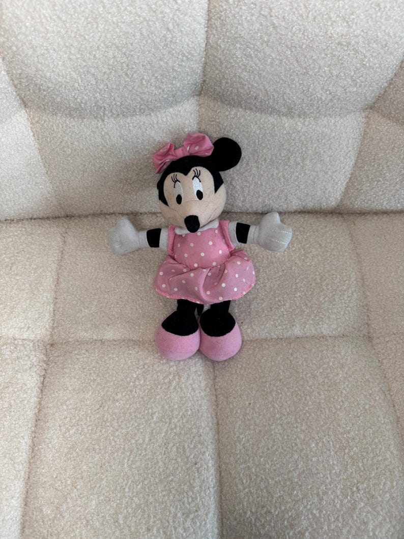 May include: A Minnie Mouse plush doll with a pink polka dot dress, matching bow, and pink shoes. The doll has black ears, a black nose, and white gloves. The plush toy is sitting on a white, textured surface.