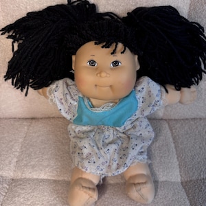 May include: A doll with a soft, light-toned face, dark brown eyes, and thick black yarn hair styled in two side ponytails. The doll wears a white dress featuring a small blue floral pattern, a solid turquoise chest panel, and white lace trim.