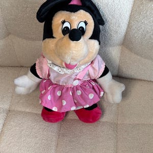 May include: A plush Minnie Mouse doll with a black head and ears, a pink bow, and a pink and white polka dot dress. The doll has white gloves and red shoes. The doll is sitting on a beige surface.