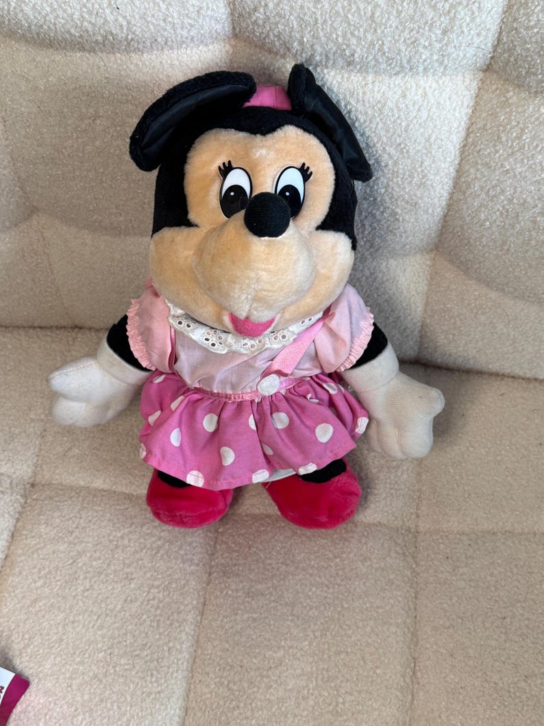 May include: A plush Minnie Mouse doll with a black head and ears, a pink bow, and a pink and white polka dot dress. The doll has white gloves and red shoes. The doll is sitting on a beige surface.