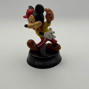 K&ouml;nnte beinhalten: Eine Mickey Mouse-Figur in Baseball-Uniform, mit roter Kappe, gelbem Hemd und roter Hose. Die Figur steht auf einem schwarzen Sockel mit der Aufschrift "SUPERSTAR".