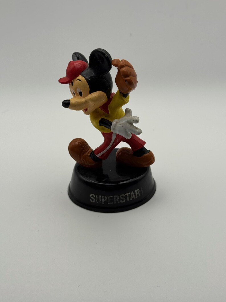 K&ouml;nnte beinhalten: Eine Mickey Mouse-Figur in Baseball-Uniform, mit roter Kappe, gelbem Hemd und roter Hose. Die Figur steht auf einem schwarzen Sockel mit der Aufschrift "SUPERSTAR".
