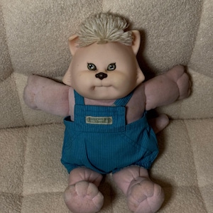 May include: A vintage Cabbage Patch Kids doll in the shape of a cat. The doll has a peach-colored face, arms, and legs, with green eyes and a tuft of light blonde hair. It wears blue overalls with a small tag.