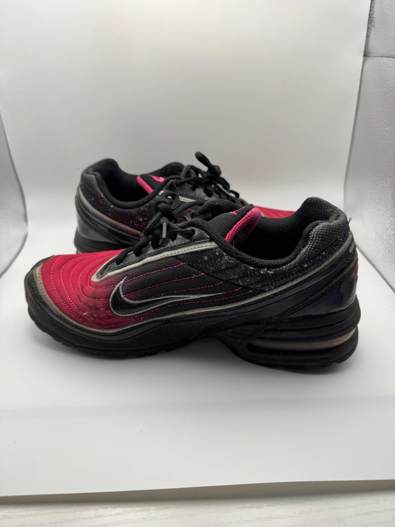 May include: Pair of black and red athletic shoes with a swoosh logo. The shoes have a black sole, black laces, and a pink accent around the ankle. The upper part of the shoe is a gradient of red to black.