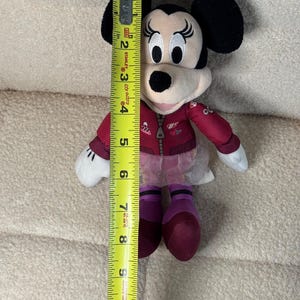 Puede incluir: Se muestra una mu&ntilde;eca de peluche de Minnie Mouse, de aproximadamente 25 cm de altura. La mu&ntilde;eca tiene orejas negras, una chaqueta roja y zapatos morados. Se sostiene una cinta m&eacute;trica amarilla junto a la mu&ntilde;eca para mostrar su altura.