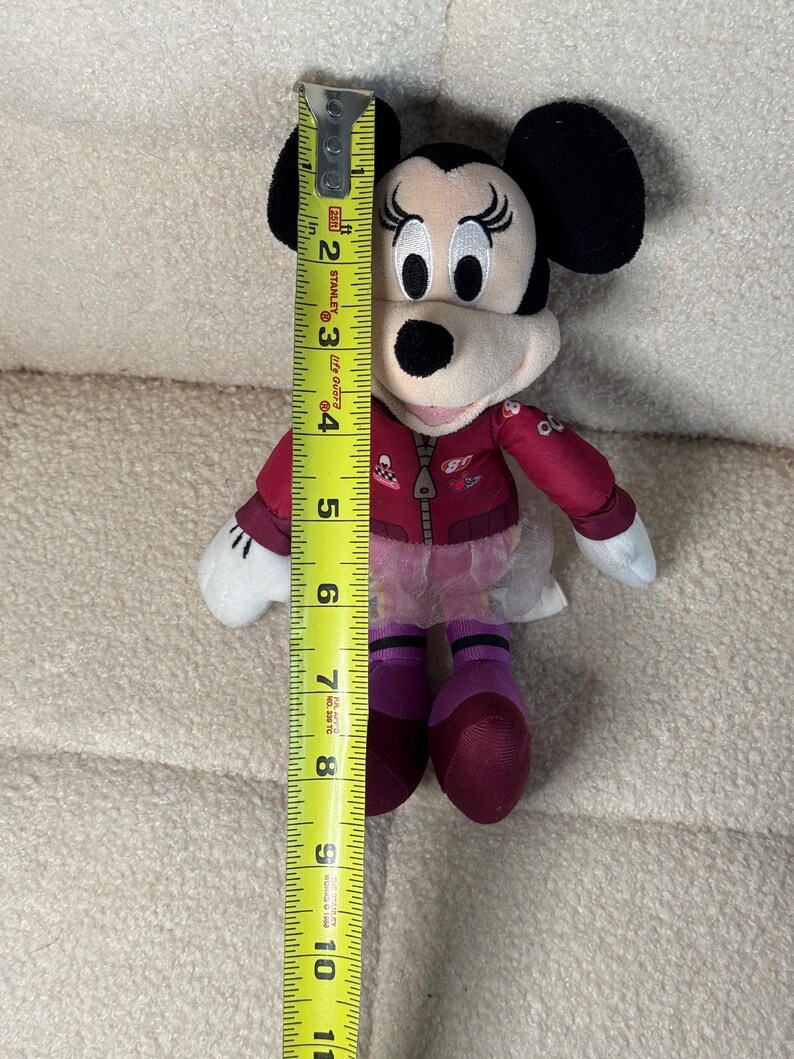 Puede incluir: Se muestra una mu&ntilde;eca de peluche de Minnie Mouse, de aproximadamente 25 cm de altura. La mu&ntilde;eca tiene orejas negras, una chaqueta roja y zapatos morados. Se sostiene una cinta m&eacute;trica amarilla junto a la mu&ntilde;eca para mostrar su altura.
