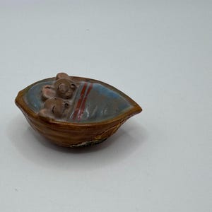 May include: A small, brown ceramic figurine of two sleeping mice nestled in a walnut shell. The shell has a blue interior with red stripes, resembling a bed. The mice are light brown with closed eyes.