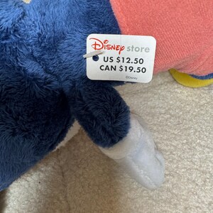May include: A soft toy in blue, pink, and white, with a Disney Store tag. The tag features the Disney Store logo and the text "US $12.50" and "CAN $19.50". The toy is made of a fluffy material.